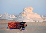A desert scene featuring a colorful patterned rug and a vehicle near white rock formations in the background.