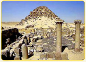 Ancient ruins with stone columns and a pyramid in the background in a desert landscape.