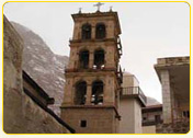 Bell tower of a historic church with multiple levels and a cross on top, set against a mountainous backdrop.