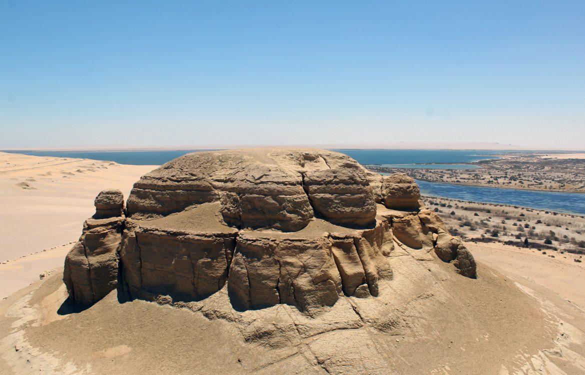 A large rock formation in a desert area with a river visible in the background.