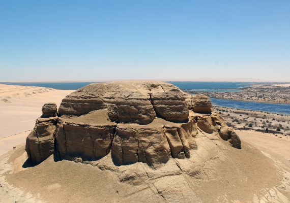 A large rock formation in a desert area with a river visible in the background.