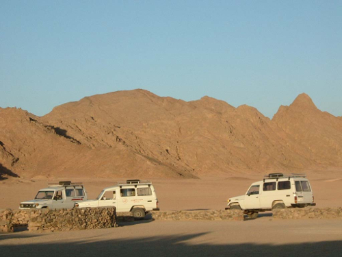 Four white SUVs parked in a desert landscape with rocky mountains in the background.