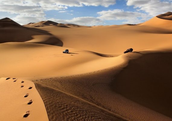 Two vehicles navigating through a vast sandy desert landscape with dunes and clouds in the sky.