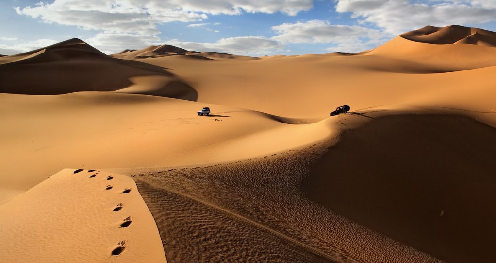 Two vehicles navigating through a vast sandy desert landscape with dunes and clouds in the sky.