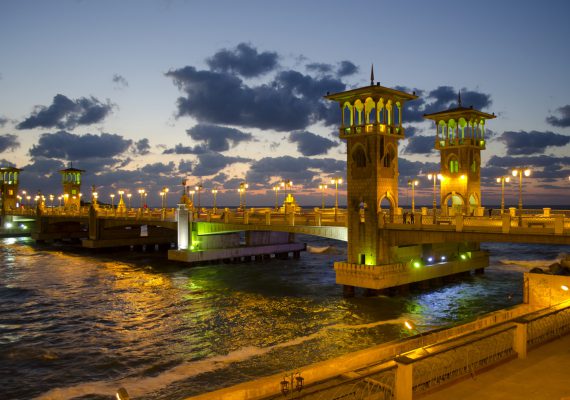 The Alexandria Corniche Bridge illuminated at sunset with clouds in the sky and water below.