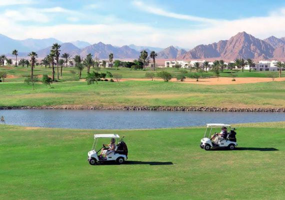 Two golf carts driving on a green golf course with mountains in the background.