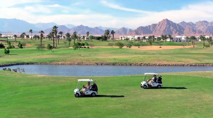 Two golf carts driving on a green golf course with mountains in the background.